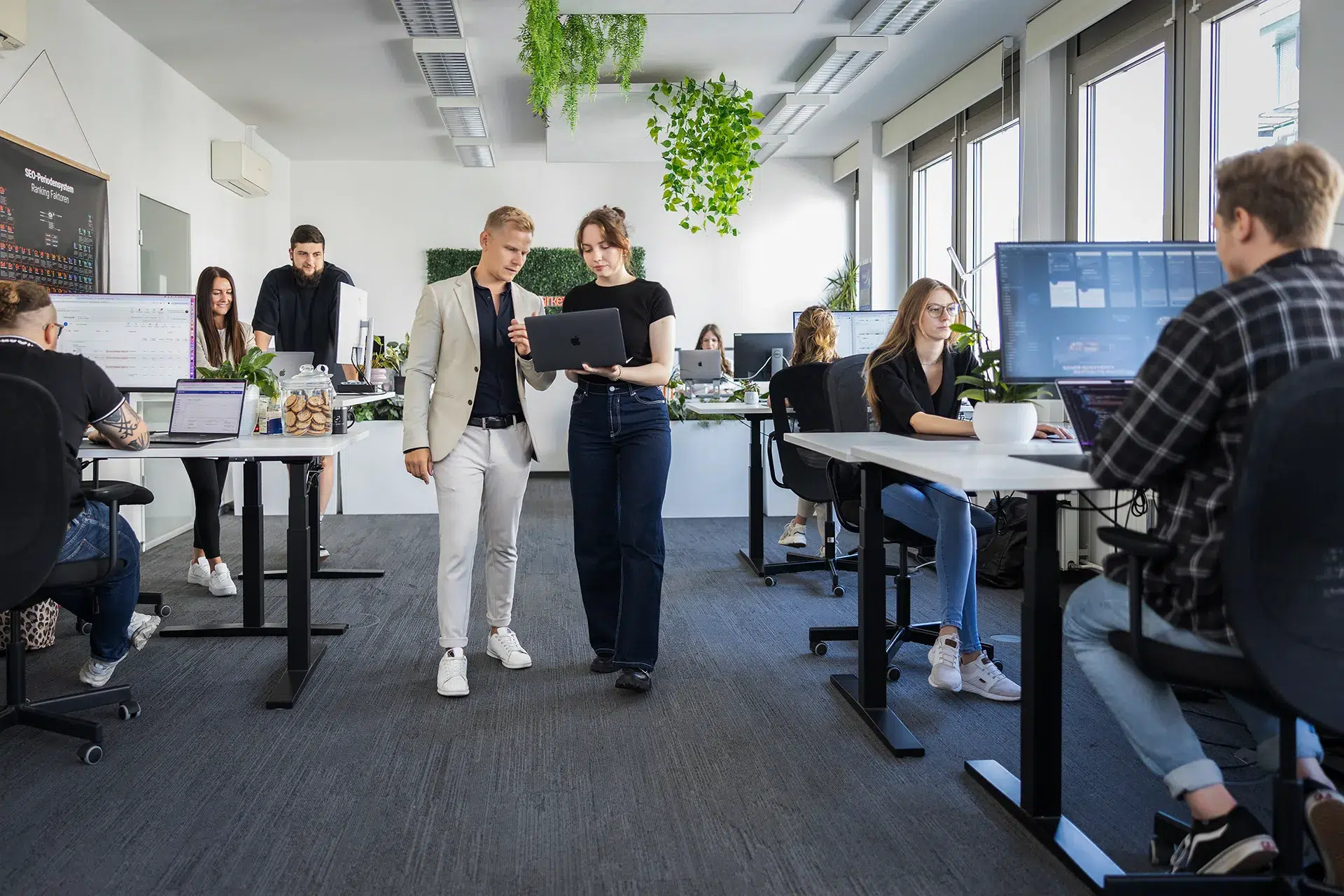 Großraumbüro mit Menschen, die an Schreibtischen an Computern arbeiten. In der Mitte laufen zwei Kollegen einer Online Marketing Agentur Dresden zusammen und besprechen etwas auf einem Tablet. Große Fenster, Zimmerpflanzen und helle Beleuchtung schaffen eine moderne Arbeitsatmosphäre.