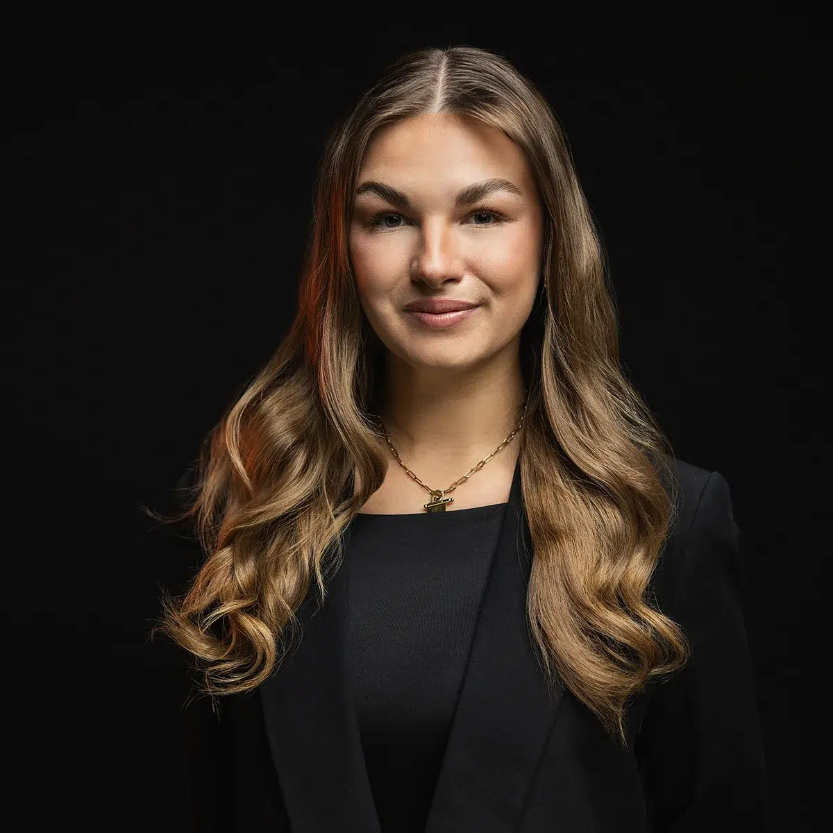 Eine Frau mit langen, welligen braunen Haaren, die einen schwarzen Blazer und eine Halskette trägt, steht vor einem schwarzen Hintergrund und lächelt sanft in die Kamera.